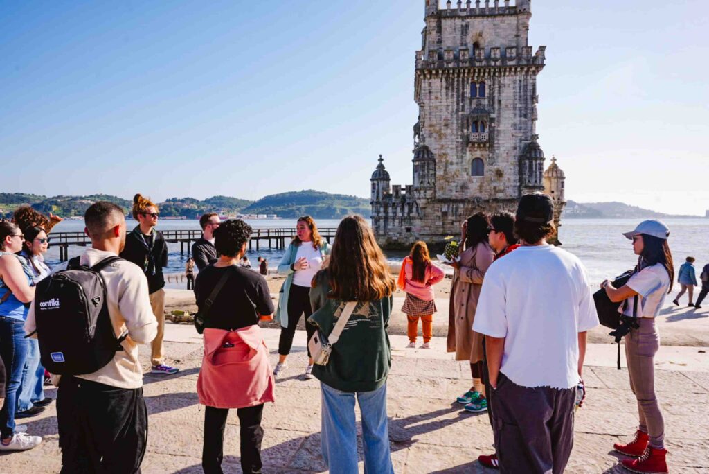 Group of travelers surrounding a trip manager tour guide who is explaining the significance of the Belem Tower in Lisbon behind them.