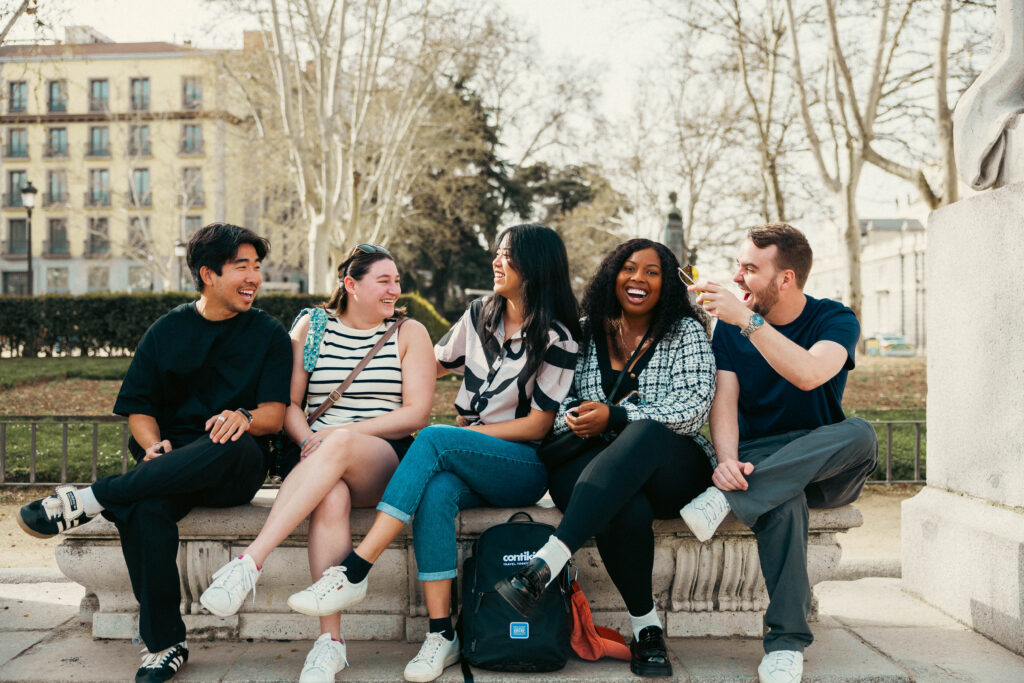Group of two men and three women friends sitting on a bench with each of their left legs crossed over their right laughing and chatting on a group trip in Madrid, Spain