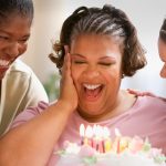 Obese woman smiles at the candles on her birthday cake as two female friends on either side of her celebrate her