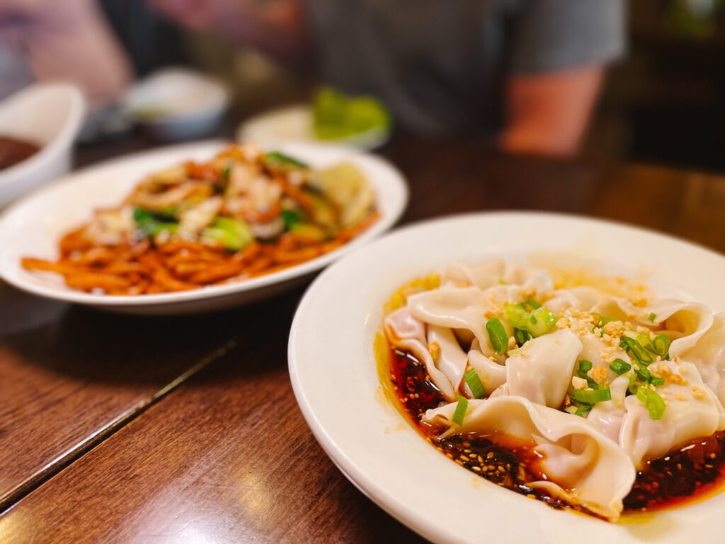 View of two Chinese family style dishes on a wooden table - one is pan fried fat noodles, the other is spicy wonton dumplings - at LJ Shanghai in Cleveland's Chinatown