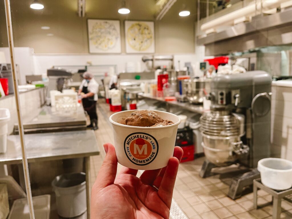Woman's outstretched hand holding a cup of chocolate peanut butter ice cream from Mitchell's in Cleveland with the ice cream shop behind where you can watch ice cream workers create the ice cream for distribution