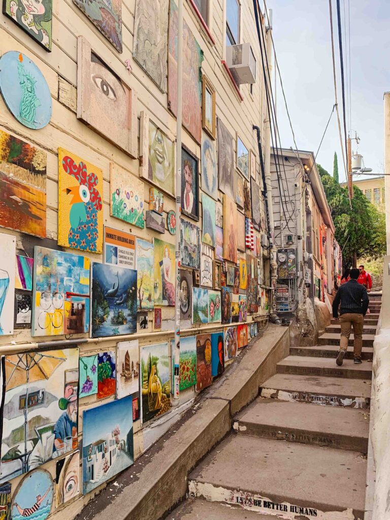 Tens of colorful paintings lining a wall adjacent to a stairway alley in Bisbee, Arizona